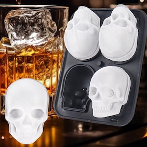 Könnte beinhalten: Eine schwarze Silikon-Eiswürfelschale mit vier Totenkopf-Formen, zusammen mit einem einzelnen Totenkopf-Eiswürfel und einem Glas bernsteinfarbener Flüssigkeit mit einem großen Eiswürfel. Die Eiswürfel sind weiß und detailliert.