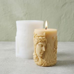 May include: A lit, beige pillar candle featuring a sculpted classical face and mountain scene. A white, cylindrical mould with embossed designs is behind the candle. The candle's flame glows against the neutral background.