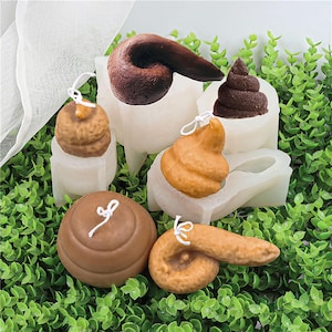 Puede incluir: Colección de velas novedosas en varias formas marrones, que se asemejan a montones de heces. Cada vela tiene una mecha blanca y está colocada en una base transparente moldeada. Las velas están dispuestas sobre césped artificial verde.