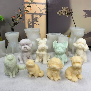 May include: A collection of nine white, beige, and green animal-shaped candles, including a cat, a poodle, a schnauzer, a bulldog, and a variety of other dog breeds. The candles are sitting on a white surface.