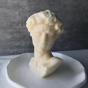 May include: A cream-coloured bust of a classical male figure, possibly a sculpture, sits on a white, organically shaped plate. The bust has detailed facial features and curly hair. The overall aesthetic is minimalist and artistic.
