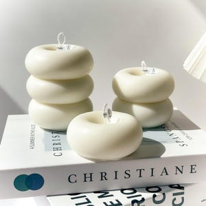 以下が含まれることがあります： 3つのアイボリー色のドーナツ型キャンドルが積み重ねられています。キャンドルには芯が付いています。「CHRISTIANE」の文字が見える白い本の上に配置されています。キャンドルはモダンなインテリアアイテムです。