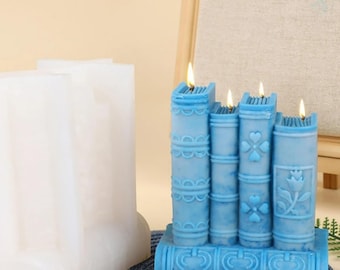 Peut inclure: Quatre bougies bleues en forme de livres, avec des mèches allumées, sont posées sur une base en forme de livre. Les bougies présentent des détails en relief. Un moule en silicone blanc du même motif de livre est visible à gauche.