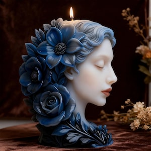 Puede incluir: Una vela decorativa con forma de cabeza de mujer, adornada con flores azules. La vela tiene una mecha encendida, y el rostro de la mujer es sereno con los ojos cerrados. Las flores incluyen rosas y otras flores detalladas, creando una pieza decorativa.