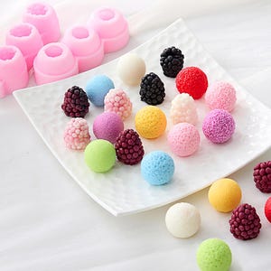 May include: A collection of colorful, spherical and berry-shaped candies and molds. The candies are in various colors, including pink, red, yellow, green, blue, white, and purple. The molds are pink and in the shape of a flower.