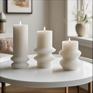 May include: Three white pillar candles with a textured, ribbed design are lit and placed on a white table. The candles vary in height and shape, with a fluted base. The flames are small and bright.