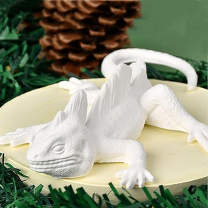 Op de afbeelding: Witte keramische hagedisfiguur met een stekelige rug en een lange staart. De hagedis ligt op zijn buik met zijn kop naar de zijkant gedraaid. De figuur staat op een witte ronde basis.