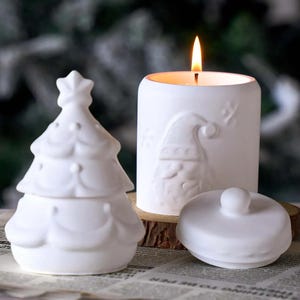 Könnte beinhalten: Ein weißer Keramik-Weihnachtsbaum-Behälter, ein weißer Kerzenhalter mit einem Weihnachtsmann-Design und eine brennende Kerze. Der Kerzenhalter befindet sich neben einem kleinen, runden, weißen Deckel.