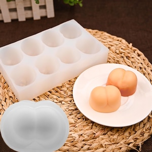 May include: A white silicone mold with eight round cavities, a white mold in the shape of a butt, and two peach-colored butt-shaped soaps on a white plate. The items are on a woven mat.