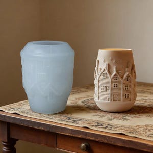 May include: A translucent light blue silicone mold featuring a raised house and building pattern sits next to a finished beige ceramic candle holder with a detailed relief of houses, chimneys, and small star shapes on its exterior surface.