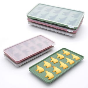 May include: A stack of three rectangular ice cube trays in green, pink, and white. Each tray has a lid and polar bear-shaped molds. One green tray holds baked bear-shaped biscuits.