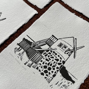 Könnte beinhalten: Eine Sammlung handgefertigter Linolschnitt-Drucke mit einem schwarz-weißen Architekturdesign. Die Drucke zeigen ein Gebäude mit strukturierter Fassade und einer windmühlenartigen Struktur. Das Papier hat einen Büttenrand, was den handwerklichen Charakter unterstreicht.
