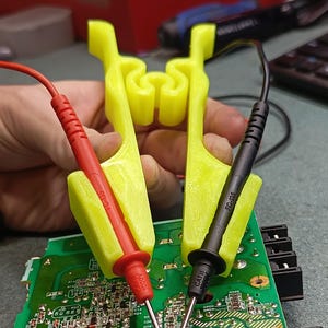 May include: A close-up of a green circuit board with red and black test probes held by yellow 3D-printed holders. The probes are connected to the board's components. The text "1000V" and "FC-135" is visible on the probes.