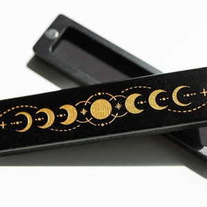 Puede incluir: Una caja de madera negra con un diseño de fases lunares doradas. La caja está abierta y el interior está forrado con fieltro negro.