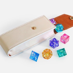 May include: A cream-coloured leather dice case with a brown suede flap, open to show several colourful polyhedral dice. The dice are various colours, including blue, pink, and orange. The case is closed with a gold snap.