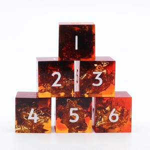 May include: A pyramid of six square resin dice with embedded gold flakes. The dice are numbered 1 through 6 in white. The resin is a gradient of orange and brown, creating a unique visual effect. The dice are arranged on a white surface.