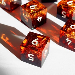 May include: Several resin dice with a fiery orange and red interior, with white numbers. The dice are cube-shaped and have a glittery, textured appearance. The dice are arranged on a white surface, with shadows.