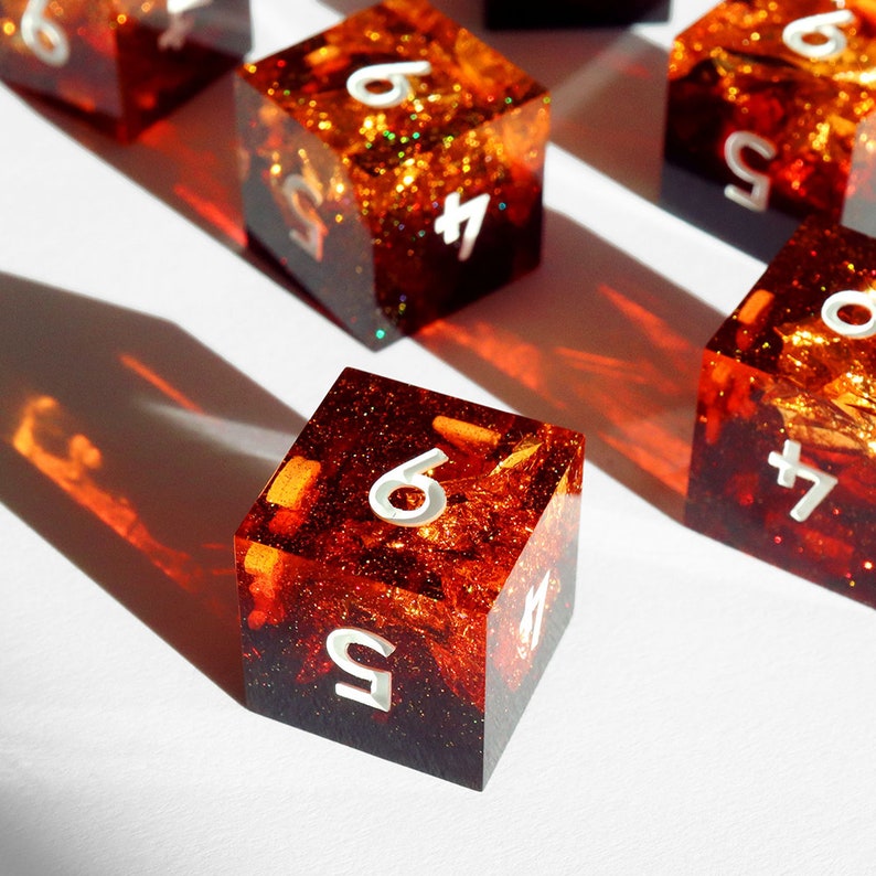 May include: Several resin dice with a fiery orange and red interior, with white numbers. The dice are cube-shaped and have a glittery, textured appearance. The dice are arranged on a white surface, with shadows.