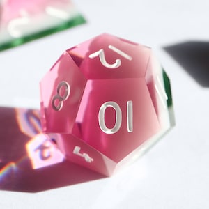 May include: A pink, clear, 10-sided dice with white numbers. The dice is made of acrylic and has a slightly frosted finish.