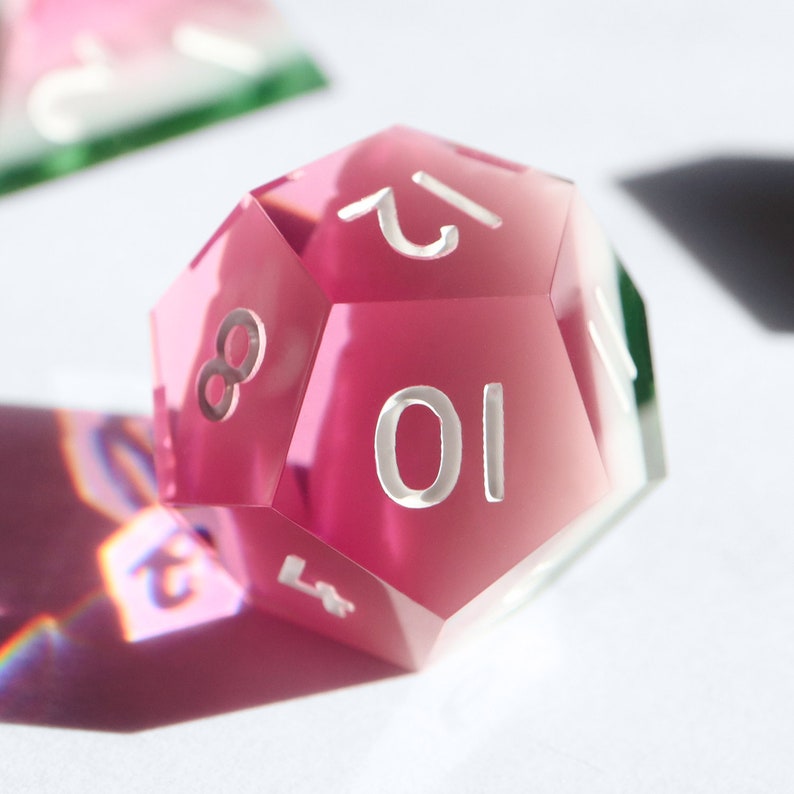 May include: A pink, clear, 10-sided dice with white numbers. The dice is made of acrylic and has a slightly frosted finish.