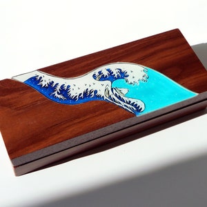 Op de afbeelding: Een houten doos met een blauw en wit golfontwerp dat op de bovenkant is geschilderd. Het golfontwerp doet denken aan het beroemde Japanse kunstwerk "De Grote Golf voor Kanagawa".