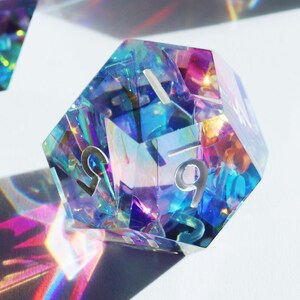 May include: A iridescent blue, purple, and pink 12-sided die with white numbers. The die is made of clear plastic and has a holographic finish.