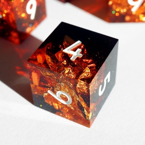 May include: A square, resin die with a fiery, gold leaf and glitter interior. The die has white, reflective numbers and a dark, smoky top. The die is a unique, handmade item for tabletop gaming.