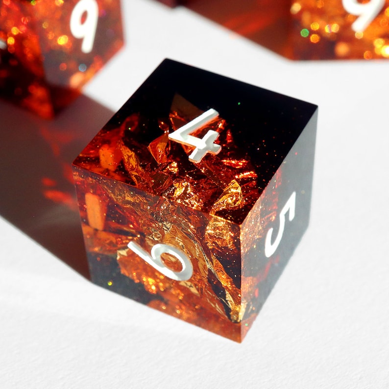 May include: A square, resin die with a fiery, gold leaf and glitter interior. The die has white, reflective numbers and a dark, smoky top. The die is a unique, handmade item for tabletop gaming.