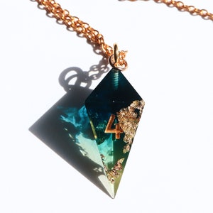 Puede incluir: Collar con colgante en forma de diamante y cadena dorada. El colgante combina resina azul y verde, pan de oro y el n&uacute;mero 4. Se muestra sobre un fondo blanco.