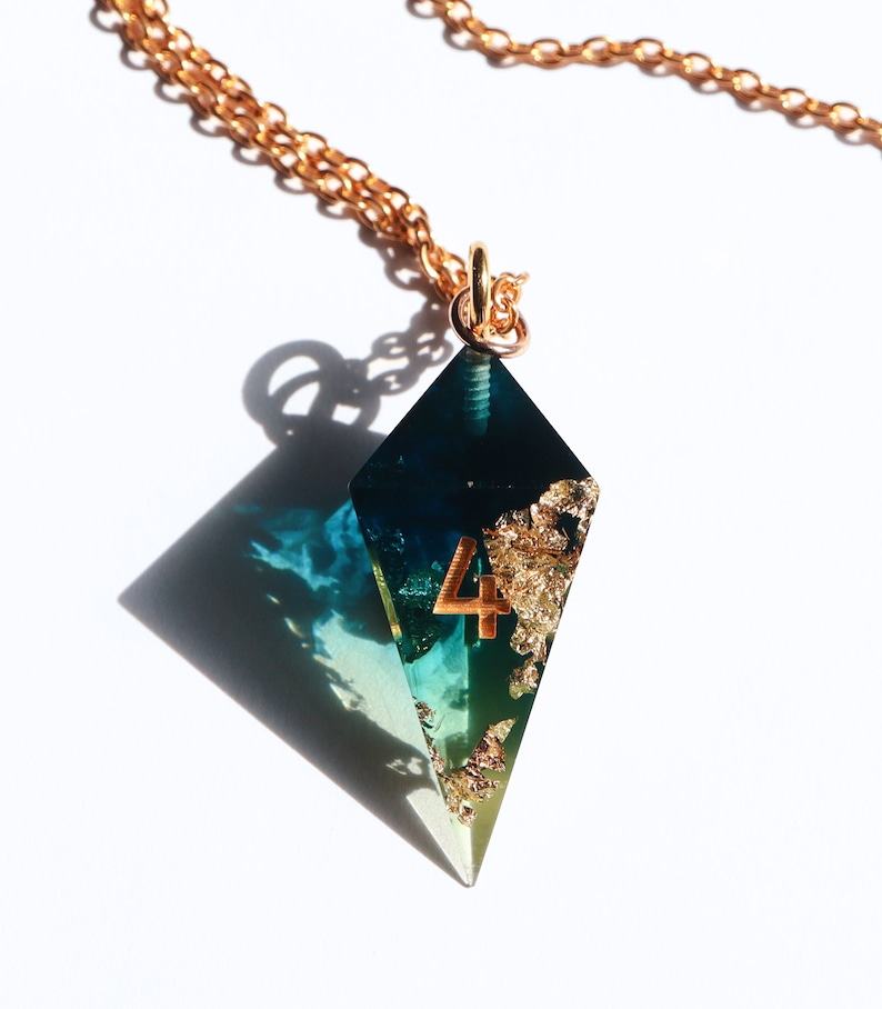 Puede incluir: Collar con colgante en forma de diamante y cadena dorada. El colgante combina resina azul y verde, pan de oro y el n&uacute;mero 4. Se muestra sobre un fondo blanco.
