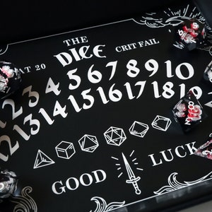May include: Black and white dice tray with the words "The Dice" and "Crit Fail" printed on it. The tray has a white outline of numbers 1-20, a sword, and the word "Luck". There are several red and white dice on the tray.