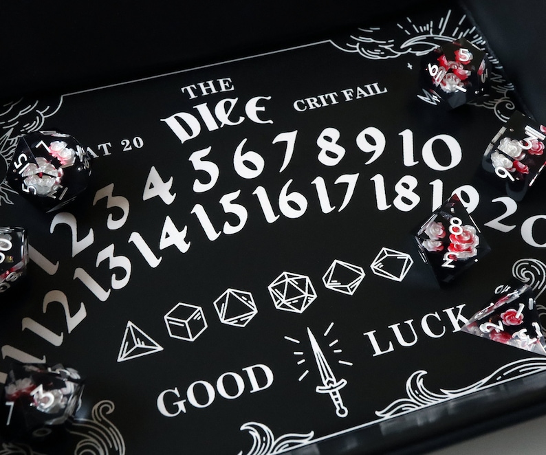 May include: Black and white dice tray with the words "The Dice" and "Crit Fail" printed on it. The tray has a white outline of numbers 1-20, a sword, and the word "Luck". There are several red and white dice on the tray.