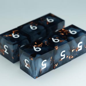 May include: Four rectangular dice with white numbers on a dark blue background with gold flecks. The dice are arranged in two rows of two, with the numbers 3, 5, and 6 visible on the faces. The dice appear to be made of resin.