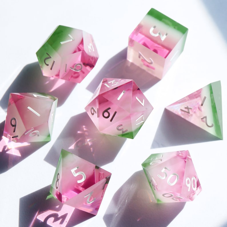 May include: Set of six pink and green dice with white numbers. The dice are made of clear acrylic and have a gradient color effect. The dice are numbered 1 through 6.