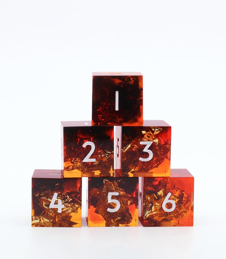 May include: A pyramid of six square resin dice with embedded gold flakes. The dice are numbered 1 through 6 in white. The resin is a gradient of orange and brown, creating a unique visual effect. The dice are arranged on a white surface.