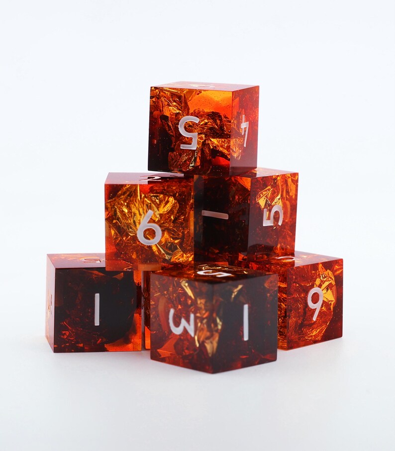 May include: A set of seven orange resin dice with gold foil inclusions. Each die has white numbers. The dice are stacked in a pyramid shape against a white background. The dice are cube-shaped.