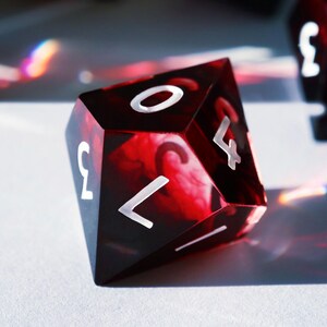 May include: A close-up of a polyhedral die with a marbled red and black design. The die has white numerals and is likely used for tabletop games. The die is resting on a white surface.
