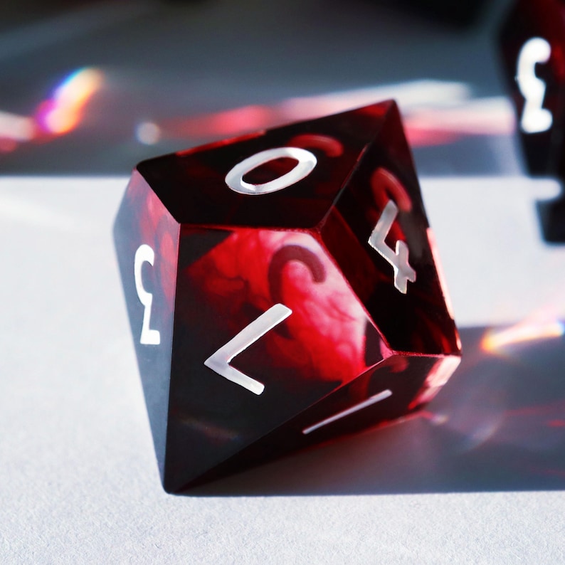 May include: A close-up of a polyhedral die with a marbled red and black design. The die has white numerals and is likely used for tabletop games. The die is resting on a white surface.