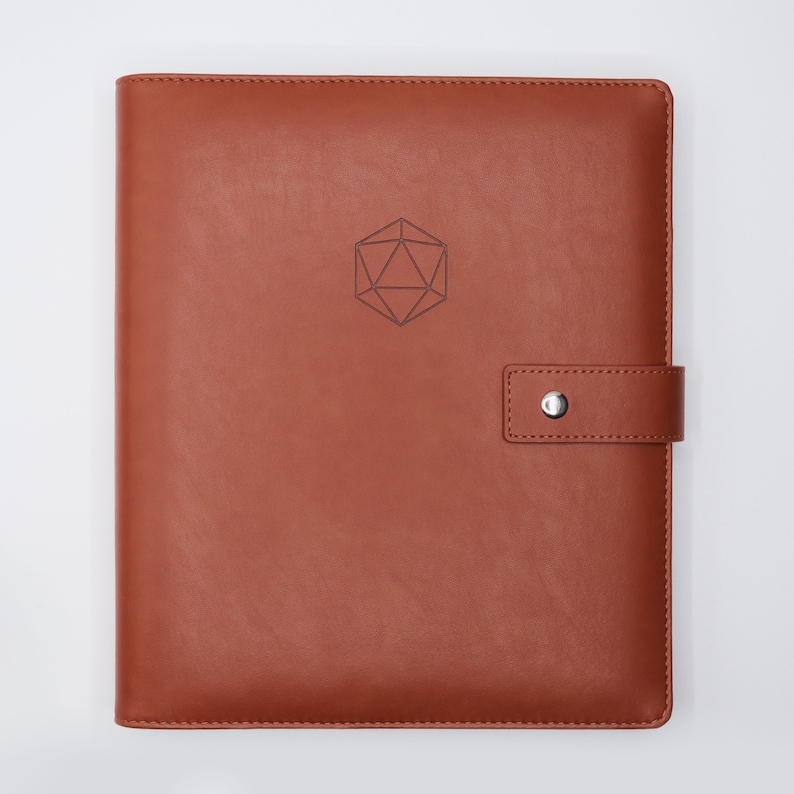 May include: A brown leather bound journal with a geometric design embossed on the front cover. The journal has a strap closure with a silver button.
