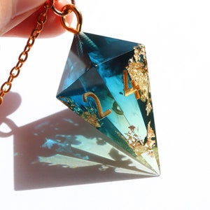 Puede incluir: Un collar de cadena dorada con un colgante geométrico grande. El colgante es de resina transparente azul-verdosa con detalles de pan de oro y los números "2" y "4" en dorado. El colgante está sostenido por una mano.