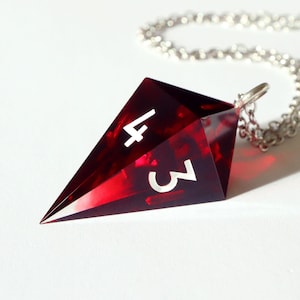 Puede incluir: Un collar con colgante geométrico rojo y negro. El colgante, con forma de dado, muestra los números 3 y 4 en blanco. Cuelga de una cadena plateada. El colgante está hecho de un material translúcido.