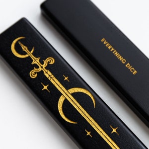 Peut inclure: Boîte en bois noir avec inscription dorée "EVERYTHING DICE". La boîte présente un motif de croissant de lune et d'épée dorés.