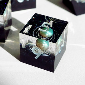 May include: A transparent resin cube with a black, glitter-filled interior. The cube displays the numbers 5 and 9 in silver. It features two teal and gold spheres with swirling designs, evoking a cosmic aesthetic.