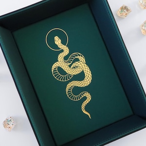 Puede incluir: Una bandeja de dados de fieltro verde con un diseño de serpiente dorada. La serpiente está enroscada y tiene un círculo alrededor de su cabeza.