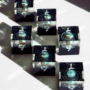 May include: Seven clear resin cubes with a black interior featuring a pearlescent orb, the number 5, and the number 9. The cubes have a cosmic design with glitter and swirling lines. The cubes are arranged on a white surface.