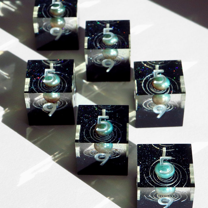 May include: Seven clear resin cubes with a black interior featuring a pearlescent orb, the number 5, and the number 9. The cubes have a cosmic design with glitter and swirling lines. The cubes are arranged on a white surface.
