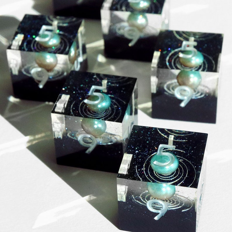 May include: A collection of clear resin cubes with a black base. Each cube displays a teal sphere, the number 5, and the number 9. The cubes have a galaxy-inspired design with glitter and swirling patterns, creating a unique aesthetic.