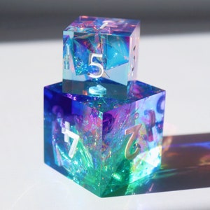 Könnte beinhalten: Zwei schimmernde Würfel, einer auf dem anderen gestapelt. Der obere Würfel ist blau und zeigt die Zahl 5. Der untere Würfel ist grün und zeigt die Zahlen 7 und 2.