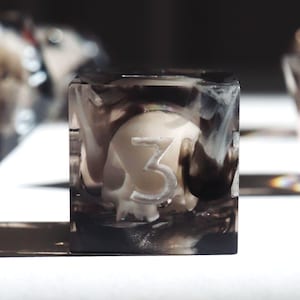 May include: A clear, square die with a skull design inside. The skull is off-white, and the number "3" is visible on one side. The die is encased in a smoky, black and white resin, creating a unique aesthetic.