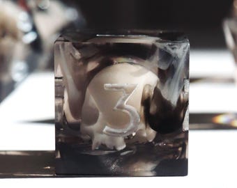 Könnte beinhalten: Ein klarer, quadratischer Würfel mit einem Totenkopf-Design im Inneren. Der Totenkopf ist cremefarben, und die Zahl "3" ist auf einer Seite sichtbar. Der Würfel ist in einem rauchigen, schwarz-weißen Harz eingeschlossen.